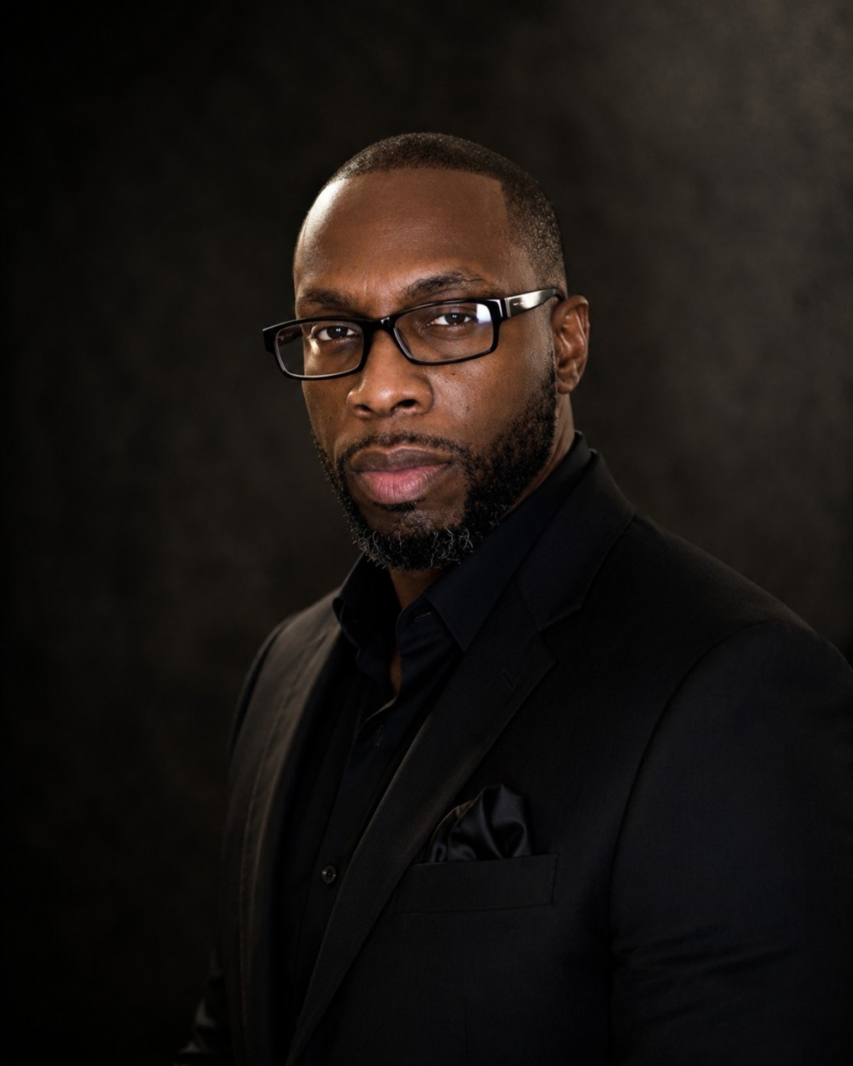 Nelly Vee official artist biography portrait wearing a black suit and black shirt against a dark studio background